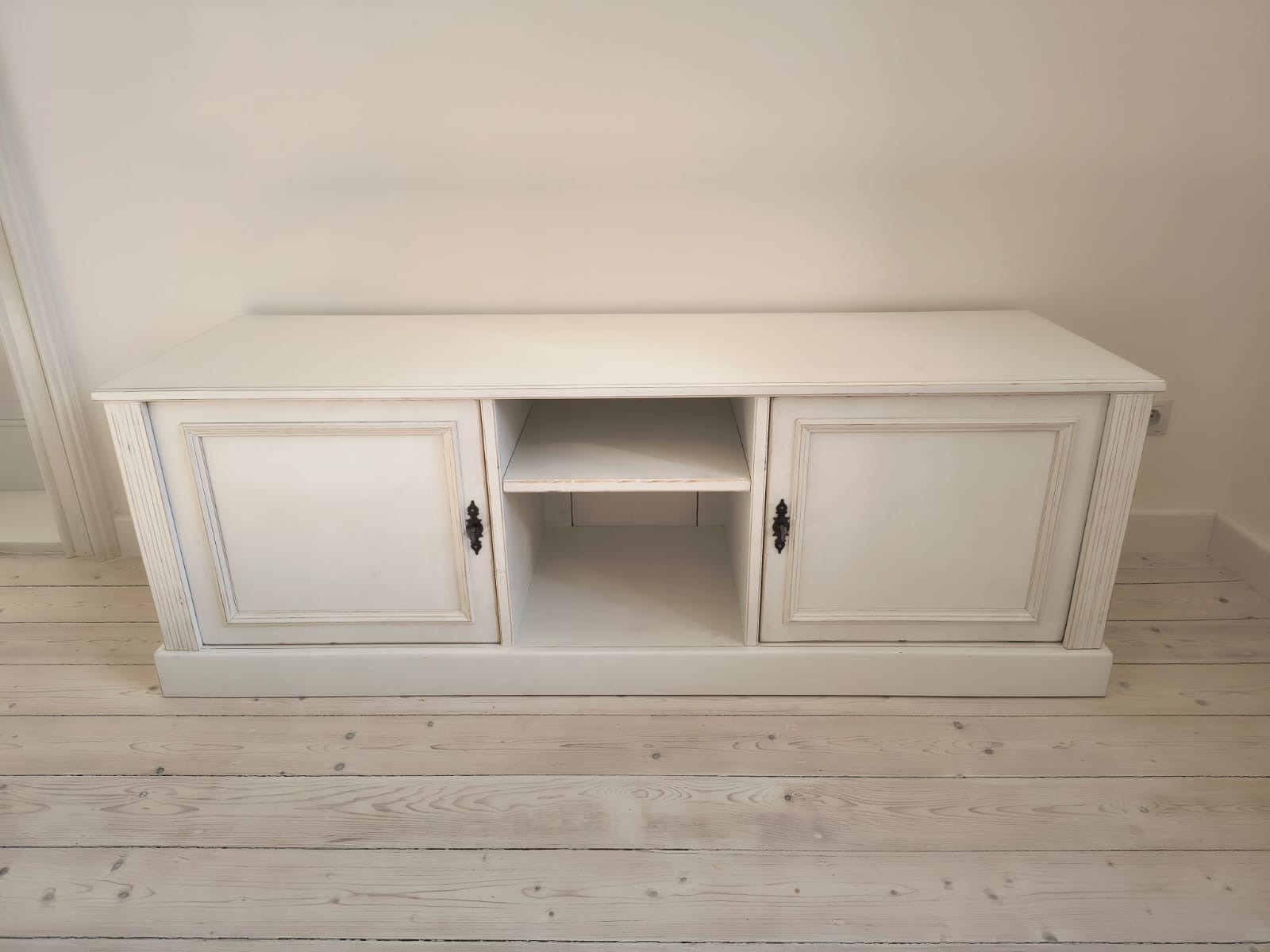 Biała, postarzana szafka RTV z dwoma zamykanymi szafkami i otwartą półką pośrodku, na jasnej drewnianej podłodze, w jasnym wnętrzu.