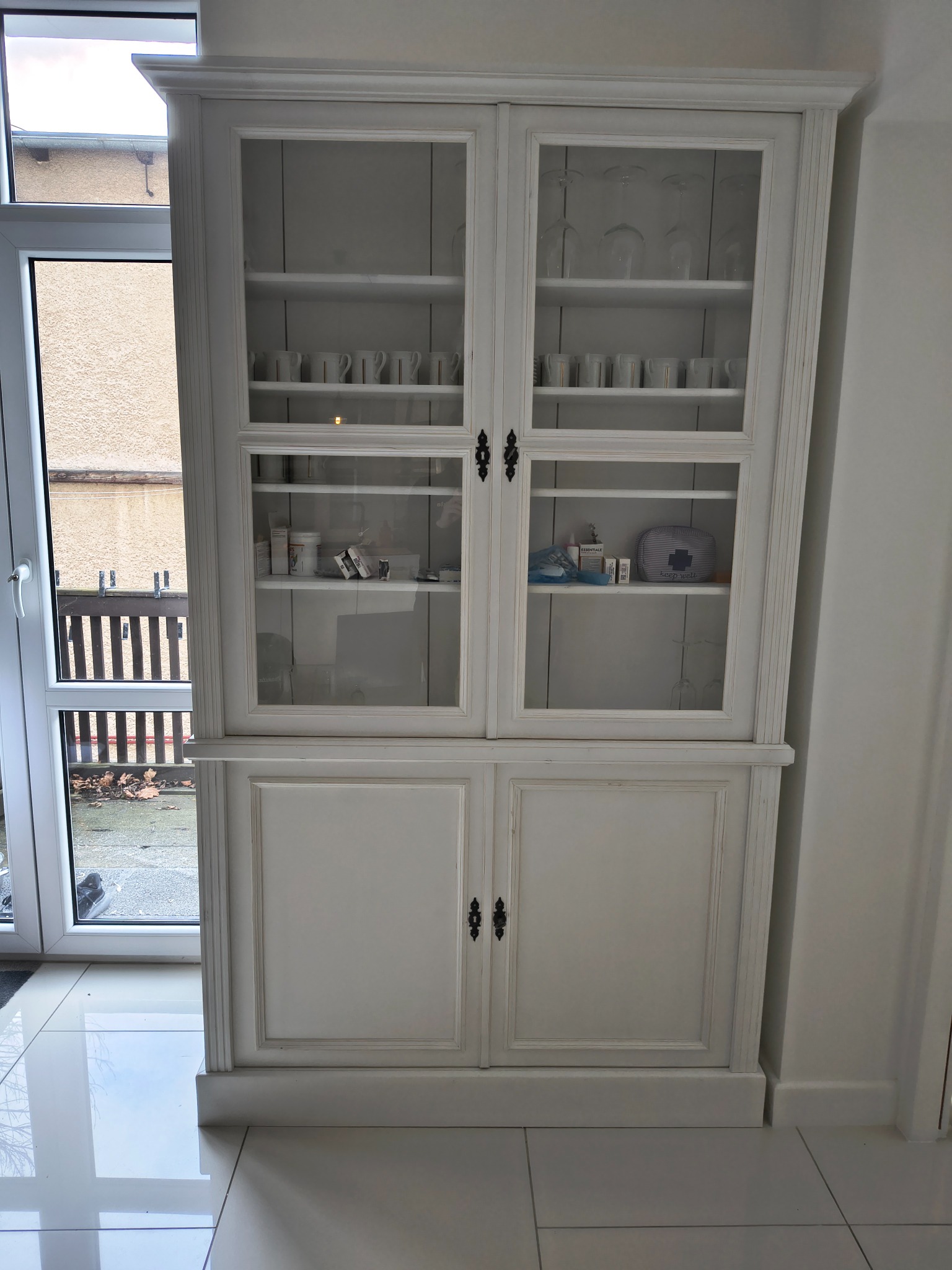 Biały, dwuczęściowy kredens z przeszkloną górą i pełnymi drzwiczkami na dole, wypełniony naczyniami i akcesoriami, ustawiony w jasnym wnętrzu obok okna.