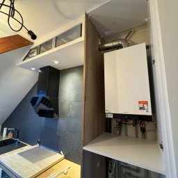 KAMIL WŁODARCZYK INSTAL KAM - Nowoczesny piec gazowy Viessmann zamontowany w szafce kuchennej, widoczne rury i instalacja. W tle czarny okap i zlewozmywak w kuchni na poddaszu.