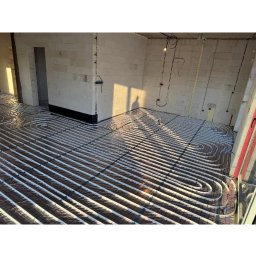 KAMIL WŁODARCZYK INSTAL KAM - Ułożone rury ogrzewania podłogowego na folii izolacyjnej w surowym wnętrzu z białego bloczka. Widoczne kable i instalacje na ścianach. W tle cień osoby.