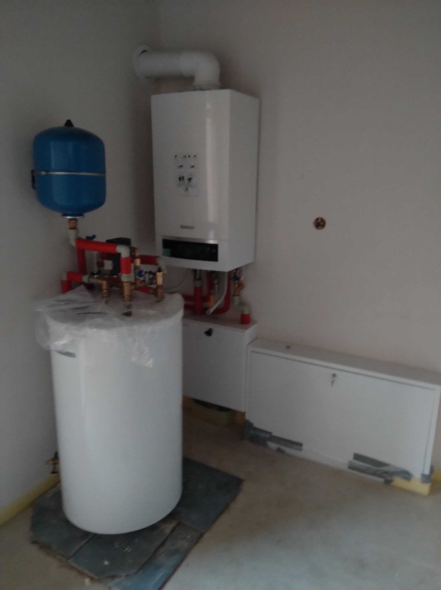 Nowoczesny piec gazowy z zbiornikiem i osprzętem w jasnym pomieszczeniu. Instalacja z rur miedzianych i tworzyw sztucznych. Zbiornik zabezpieczony folią.