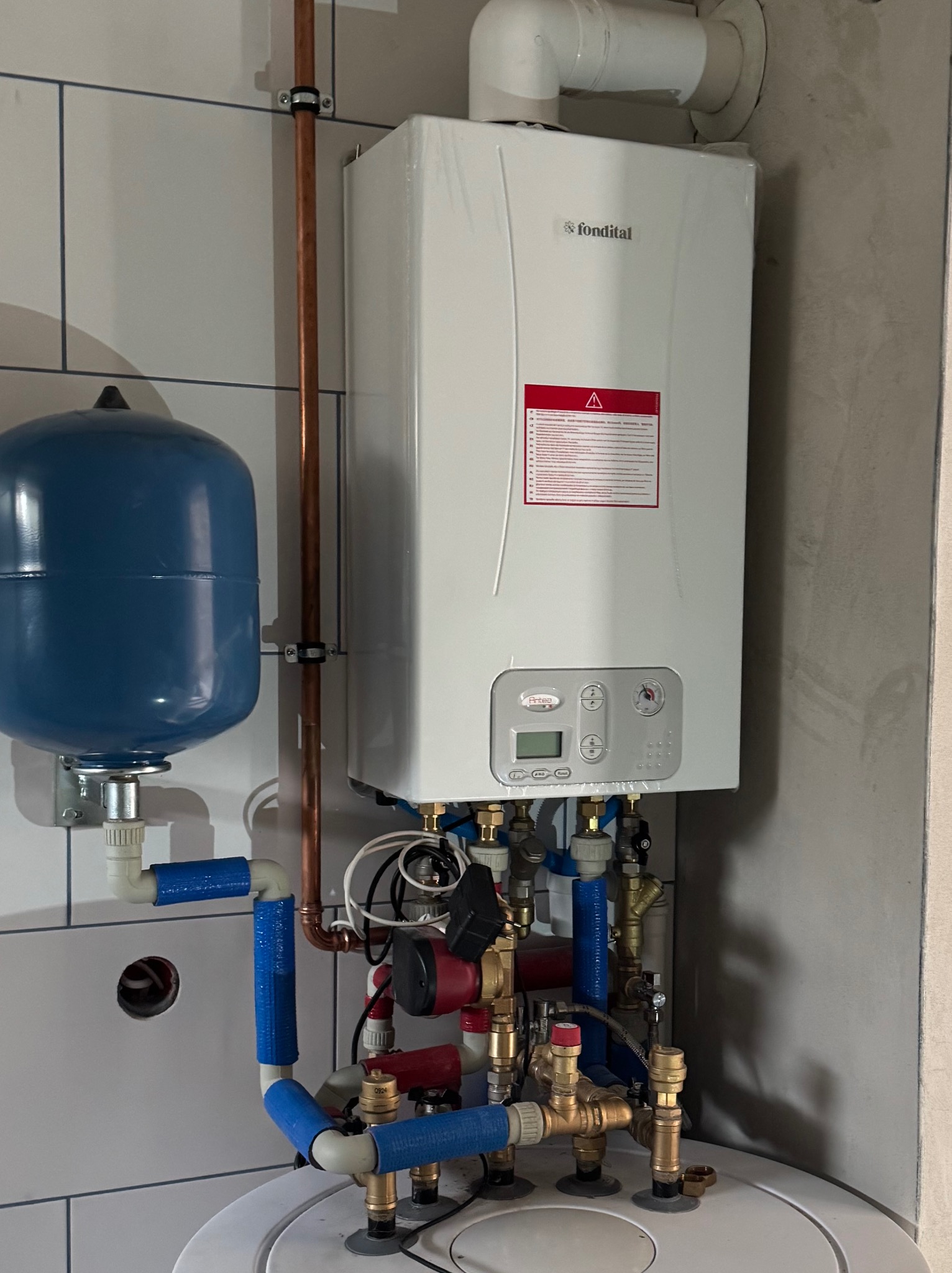 Biały piec gazowy Fondital z niebieskim zbiornikiem wyrównawczym i miedzianymi rurami na tle białych płytek i surowej ściany. Instalacja hydrauliczna.