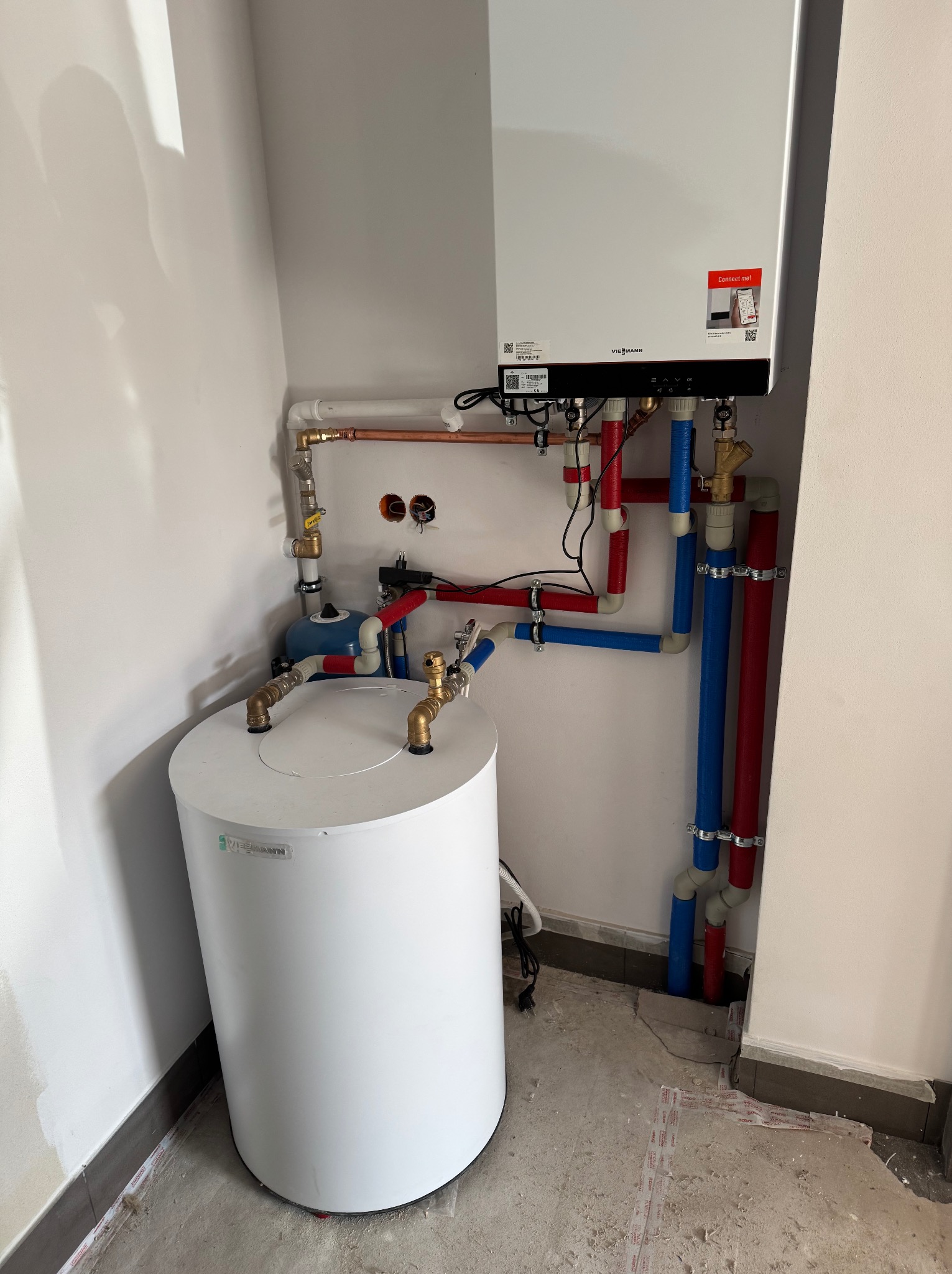 Nowoczesny piec gazowy Viessmann z podłączonym zasobnikiem ciepłej wody i instalacją z rur w kolorach czerwonym i niebieskim w narożniku pomieszczenia.