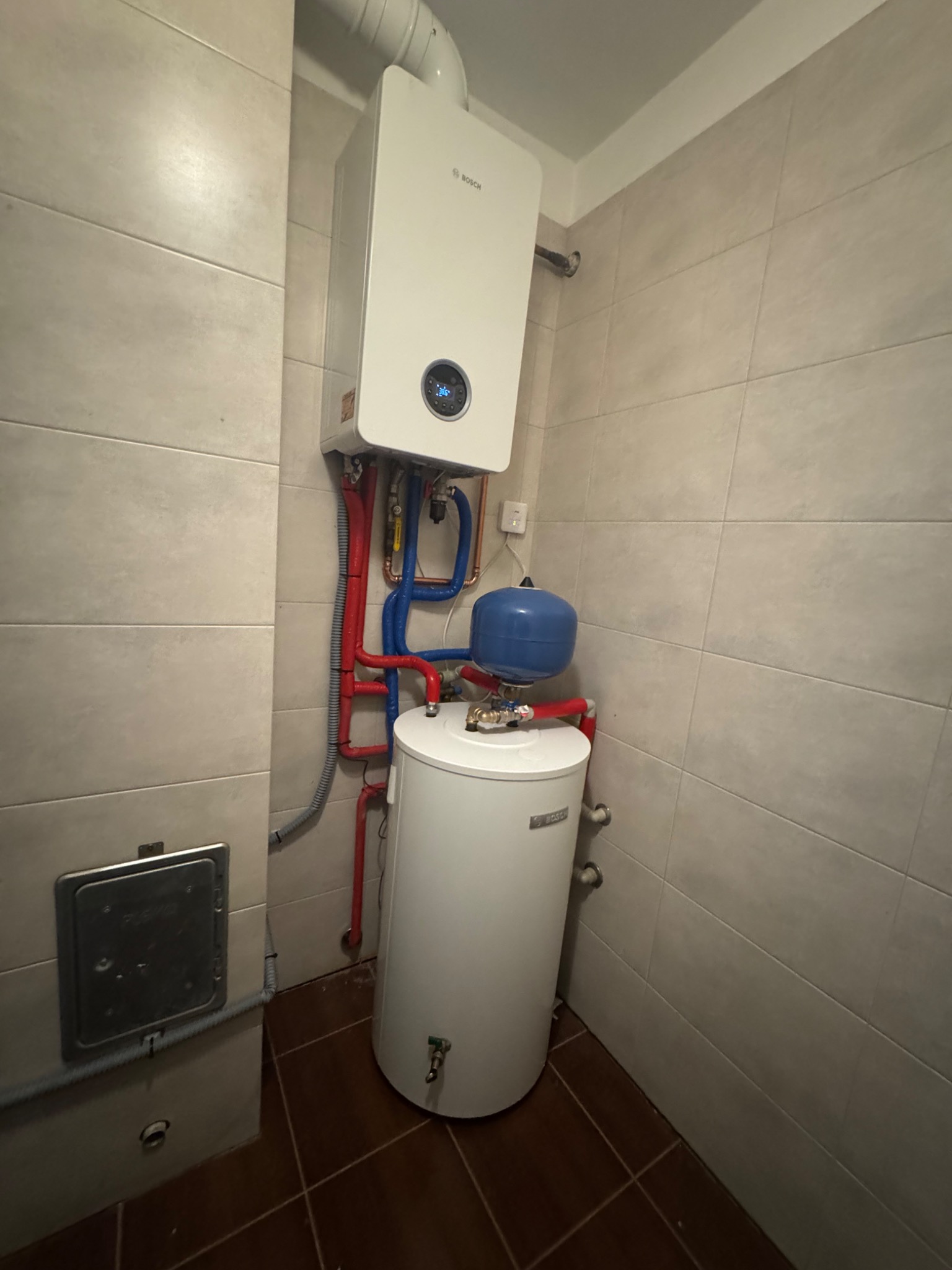 Biały piec gazowy Bosch z podłączonym zasobnikiem wody i instalacją w kolorze czerwonym i niebieskim, na tle beżowych płytek w pomieszczeniu.