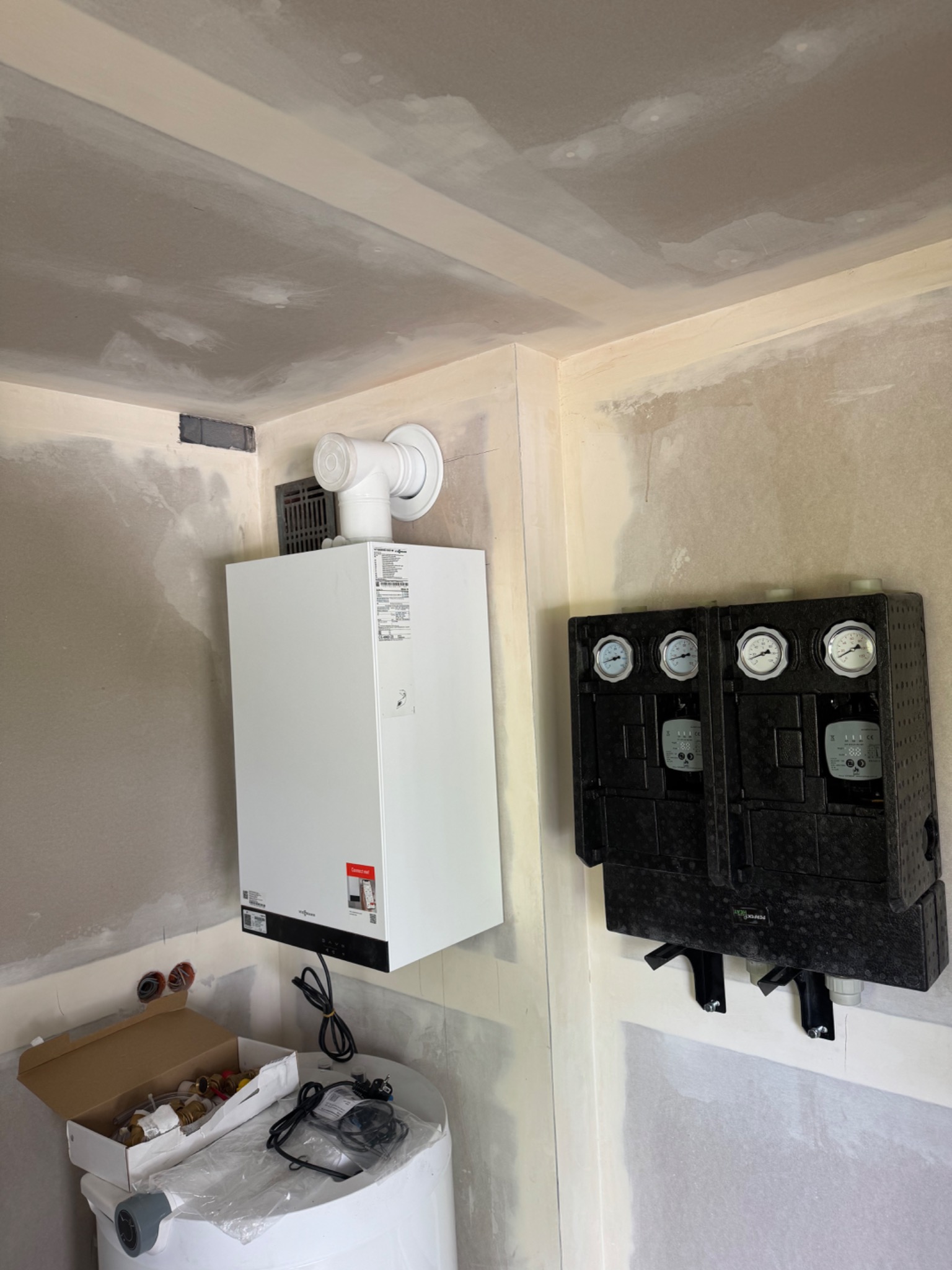 Biały piec gazowy Viessmann zamontowany na ścianie w surowym pomieszczeniu, obok czarny rozdzielacz z manometrami. Widoczne akcesoria instalacyjne.