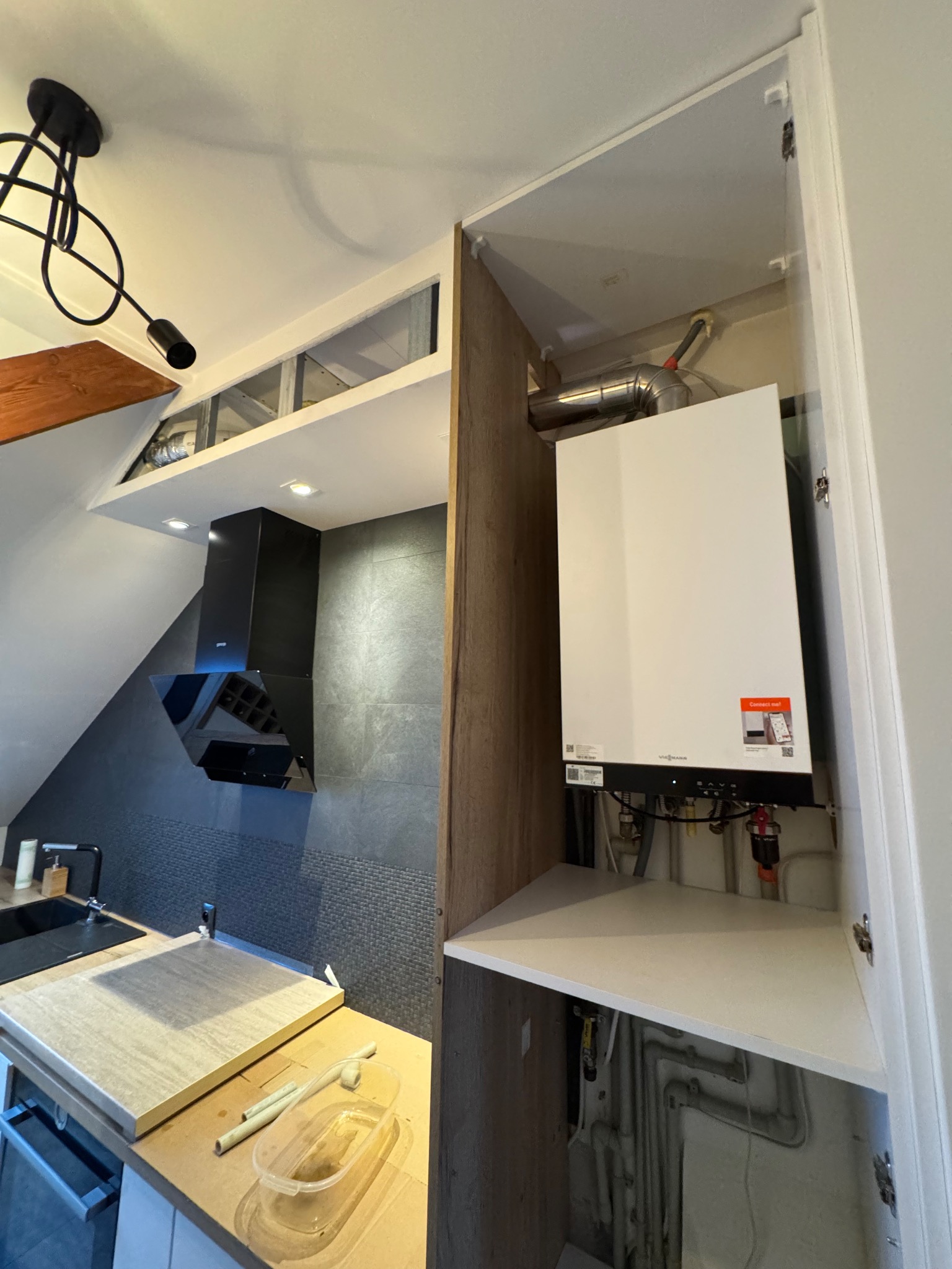 Nowoczesny piec gazowy Viessmann zamontowany w szafce kuchennej, widoczne rury i instalacja. W tle czarny okap i zlewozmywak w kuchni na poddaszu.