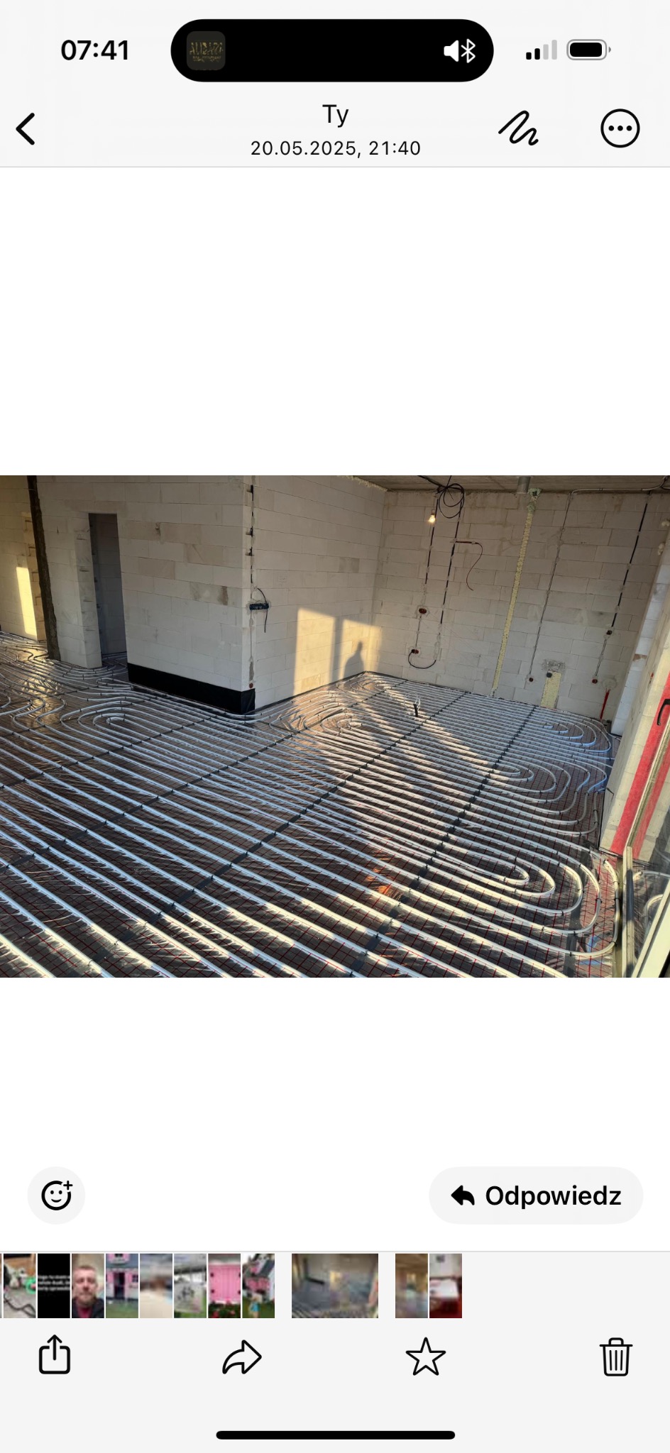 Ułożone rury ogrzewania podłogowego na folii izolacyjnej w surowym wnętrzu z białego bloczka. Widoczne kable i instalacje na ścianach. W tle cień osoby.