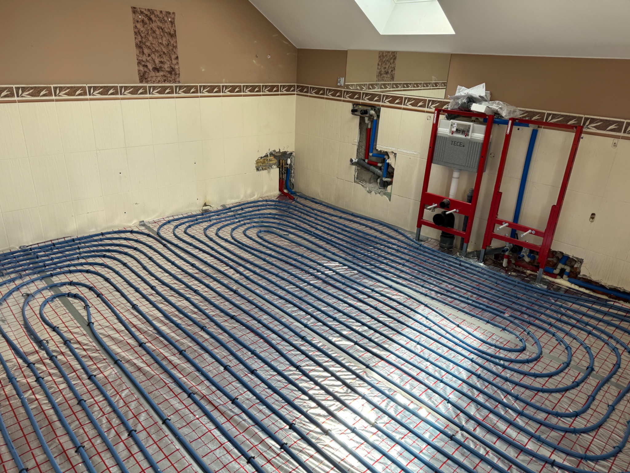 Niebieskie rury ogrzewania podłogowego ułożone na folii z czerwoną siatką w łazience z częściowo wykończonymi ścianami i stelażem podtynkowym WC.