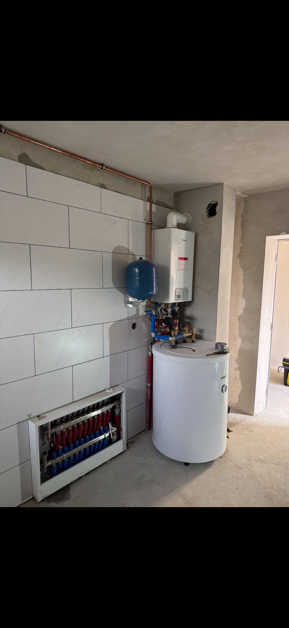 Zmontowana instalacja grzewcza z piecem gazowym, zasobnikiem CWU i rozdzielaczem ogrzewania podłogowego w surowym pomieszczeniu z wykończoną ścianą z płytek.