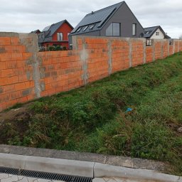 Usługi ogólnobudowlane i remontowe - Wznoszenie muru ogrodzeniowego z czerwonej cegły dziurawki, widoczne słupki betonowe, w tle nowoczesne domy z panelami słonecznymi na dachu, pochmurne niebo.