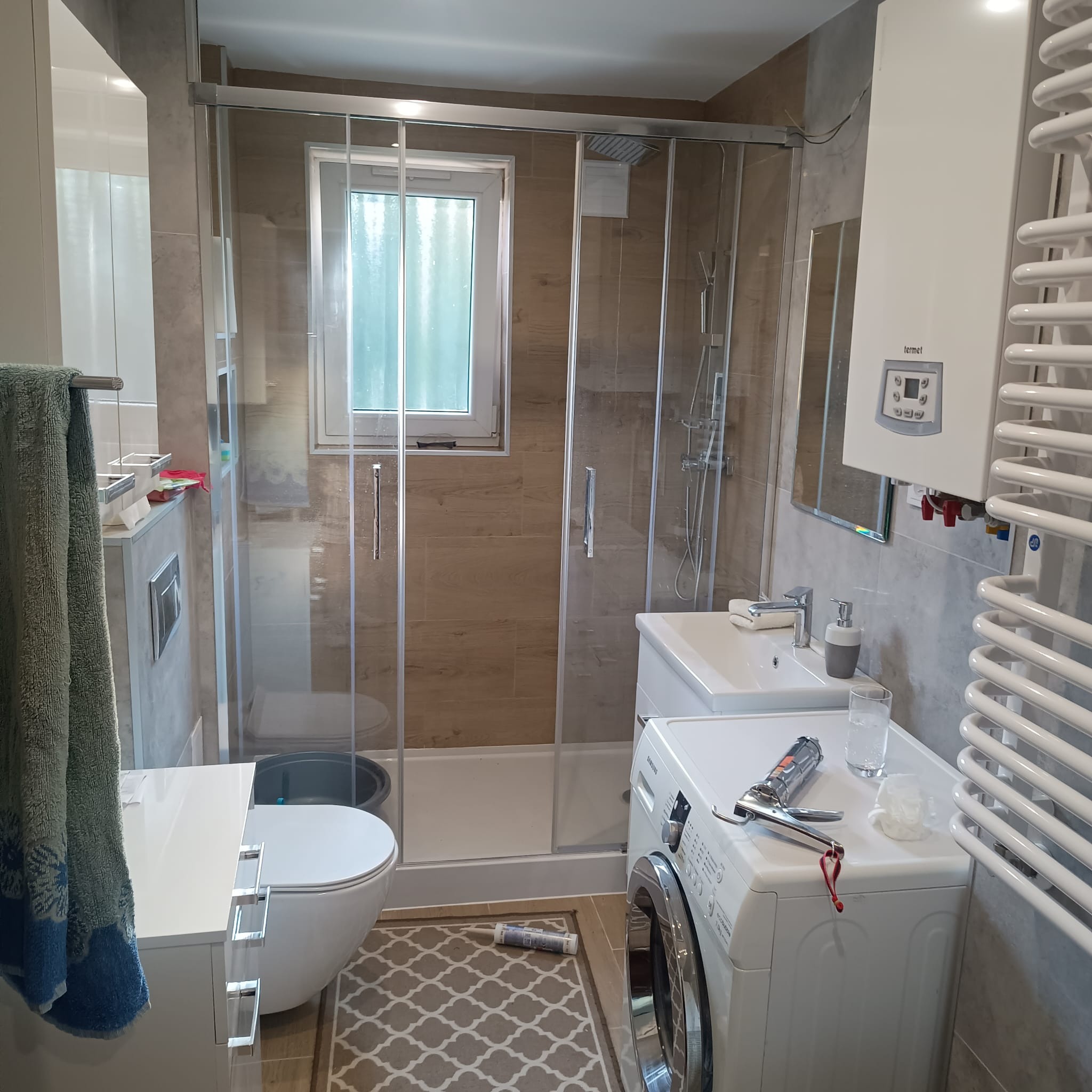 Wykończona łazienka z kabiną prysznicową, umywalką, pralką, grzejnikiem drabinkowym i toaletą, na pralce pistolet do silikonu i szklanka z wodą, na podłodze dywanik i tuba silikonu.