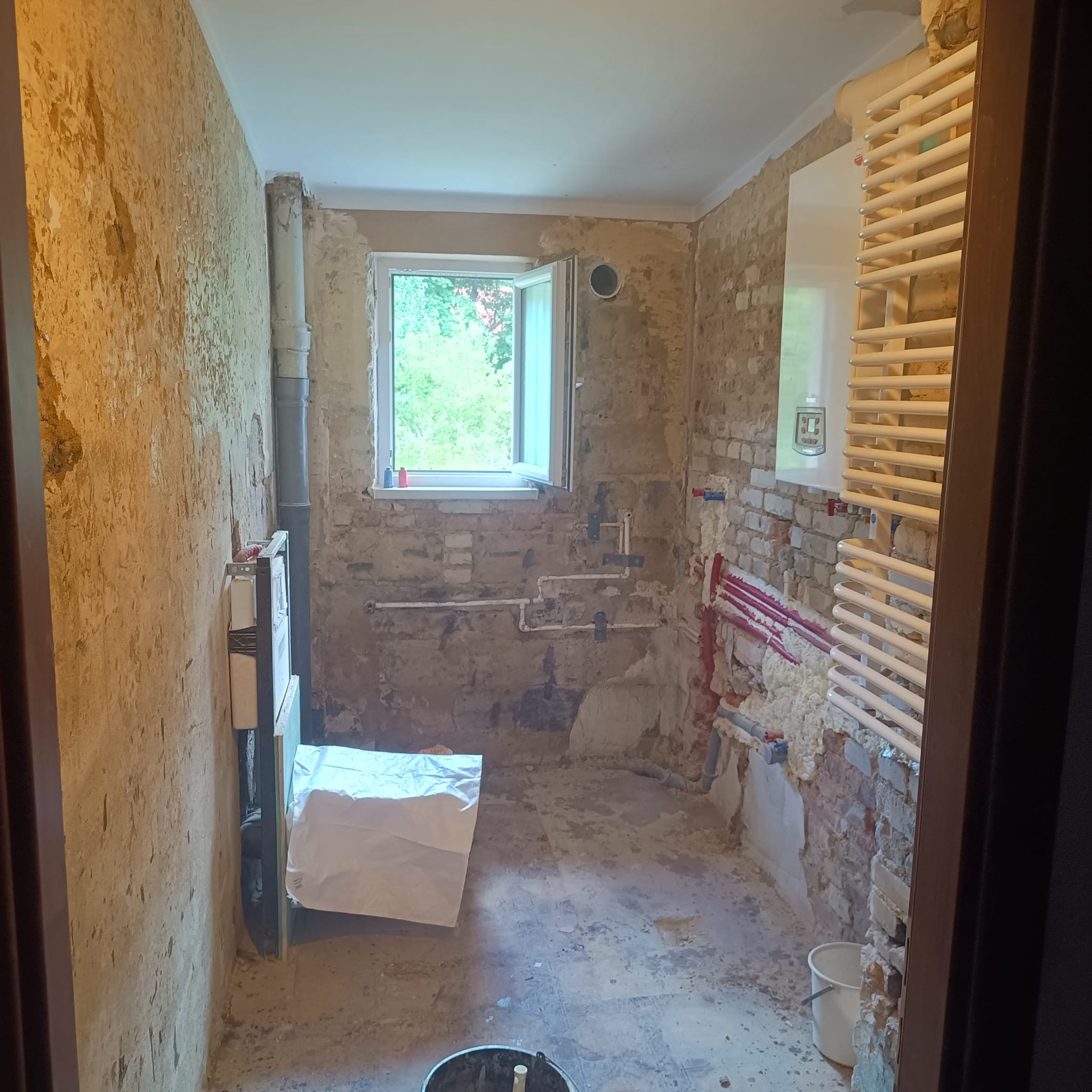 Łazienka w trakcie remontu: odsłonięta cegła, nowe rury, grzejnik drabinkowy, okno z widokiem na zieleń, ściany przygotowane do tynkowania.