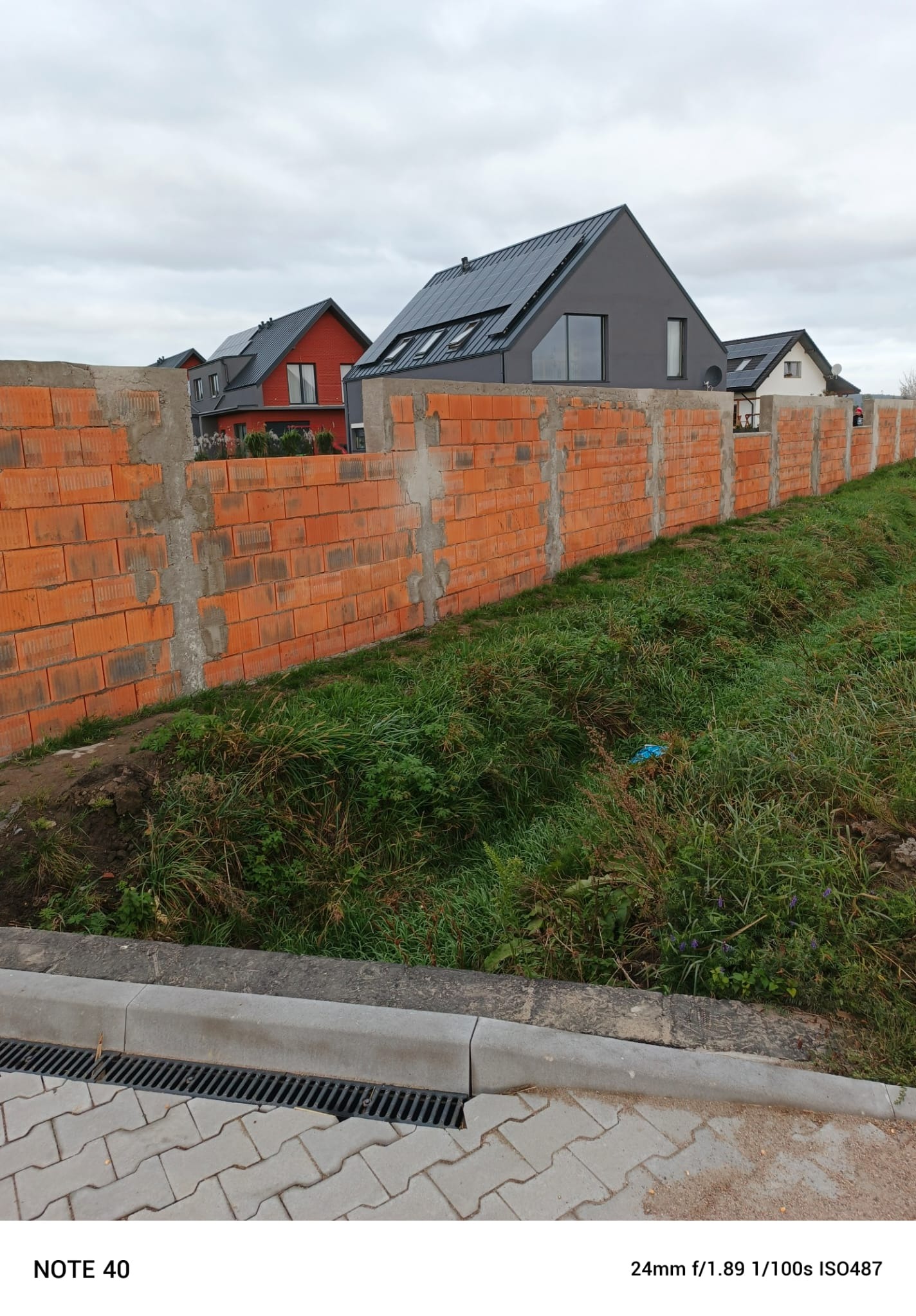 Wznoszenie muru ogrodzeniowego z czerwonej cegły dziurawki, widoczne słupki betonowe, w tle nowoczesne domy z panelami słonecznymi na dachu, pochmurne niebo.