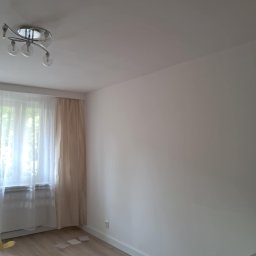 Oskar Fundalski - Jasne wnętrze po remoncie: świeżo pomalowana ściana, panele podłogowe w odcieniach drewna, okno z firankami i nowoczesna lampa sufitowa. Minimalistyczny design.