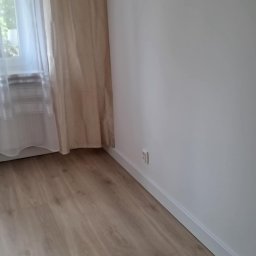 Oskar Fundalski - Jasne panele podłogowe w pokoju z beżową zasłoną przy oknie i białą ścianą z listwą przypodłogową. Widoczny fragment gniazdka elektrycznego.