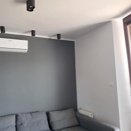 Oskar Fundalski - Pokój po malowaniu z szarą ścianą, klimatyzacją i nowoczesnym oświetleniem sufitowym. Widoczny narożnik w odcieniach szarości. Minimalistyczny design wnętrza.