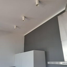 Oskar Fundalski - Nowoczesna kuchnia po malowaniu: szara ściana kontrastuje z białymi szafkami i sufitowymi lampami tubami. Widoczny fragment okapu kuchennego.