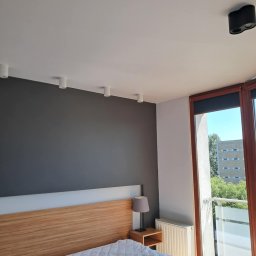 Oskar Fundalski - Jasne wnętrze sypialni z szarą ścianą, drewnianym zagłówkiem i oknem balkonowym z widokiem na zieleń. Oświetlenie sufitowe punktowe.