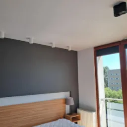 Jasne wnętrze sypialni z szarą ścianą, drewnianym zagłówkiem i oknem balkonowym z widokiem na zieleń. Oświetlenie sufitowe punktowe.