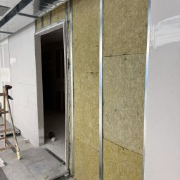 Oskar Fundalski - Montaż izolacji z wełny mineralnej w metalowym stelażu ściany działowej, widoczne płyty gipsowo-kartonowe i drabina. Wnętrze w trakcie prac wykończeniowych.