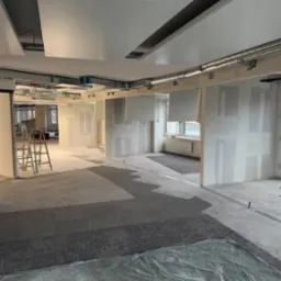 Biuro w trakcie remontu: widoczne ściany z płyt karton-gips, metalowe rury pod sufitem, drabina i zabezpieczona podłoga. Etap prac wykończeniowych, surowy stan.