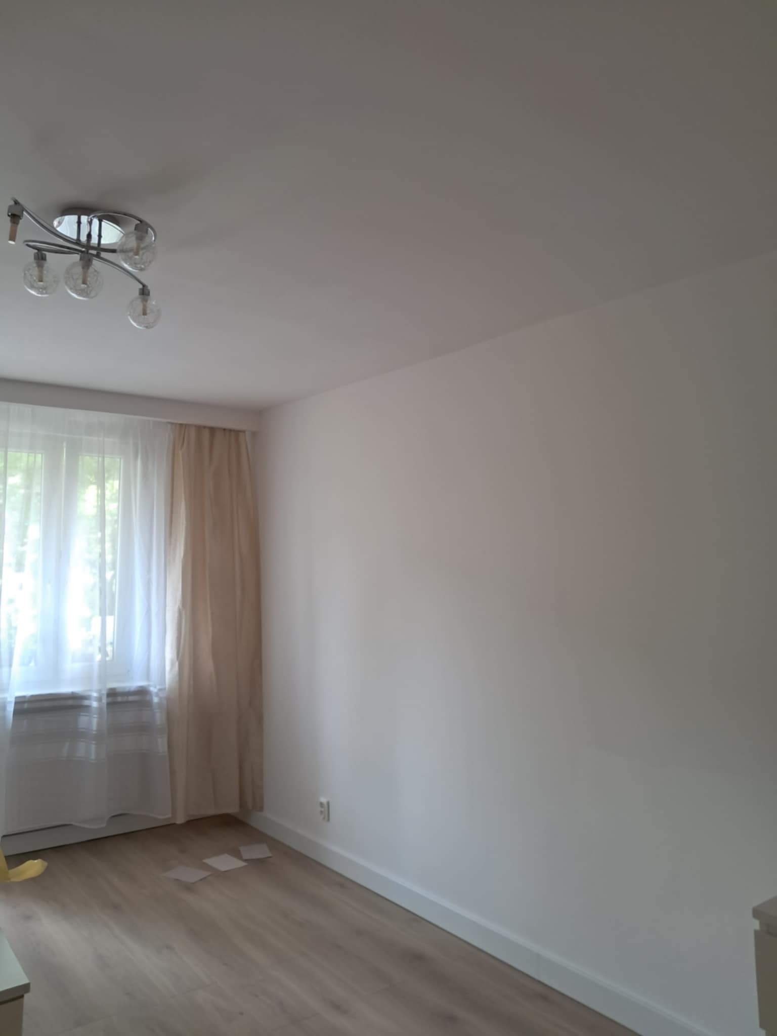 Jasne wnętrze po remoncie: świeżo pomalowana ściana, panele podłogowe w odcieniach drewna, okno z firankami i nowoczesna lampa sufitowa. Minimalistyczny design.