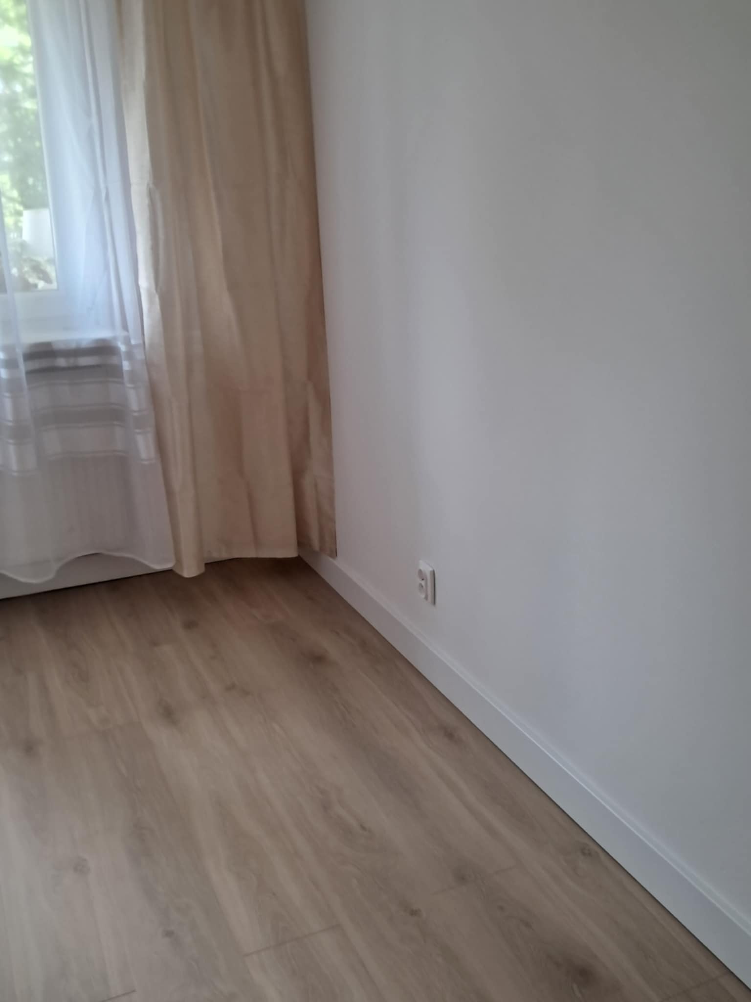 Jasne panele podłogowe w pokoju z beżową zasłoną przy oknie i białą ścianą z listwą przypodłogową. Widoczny fragment gniazdka elektrycznego.