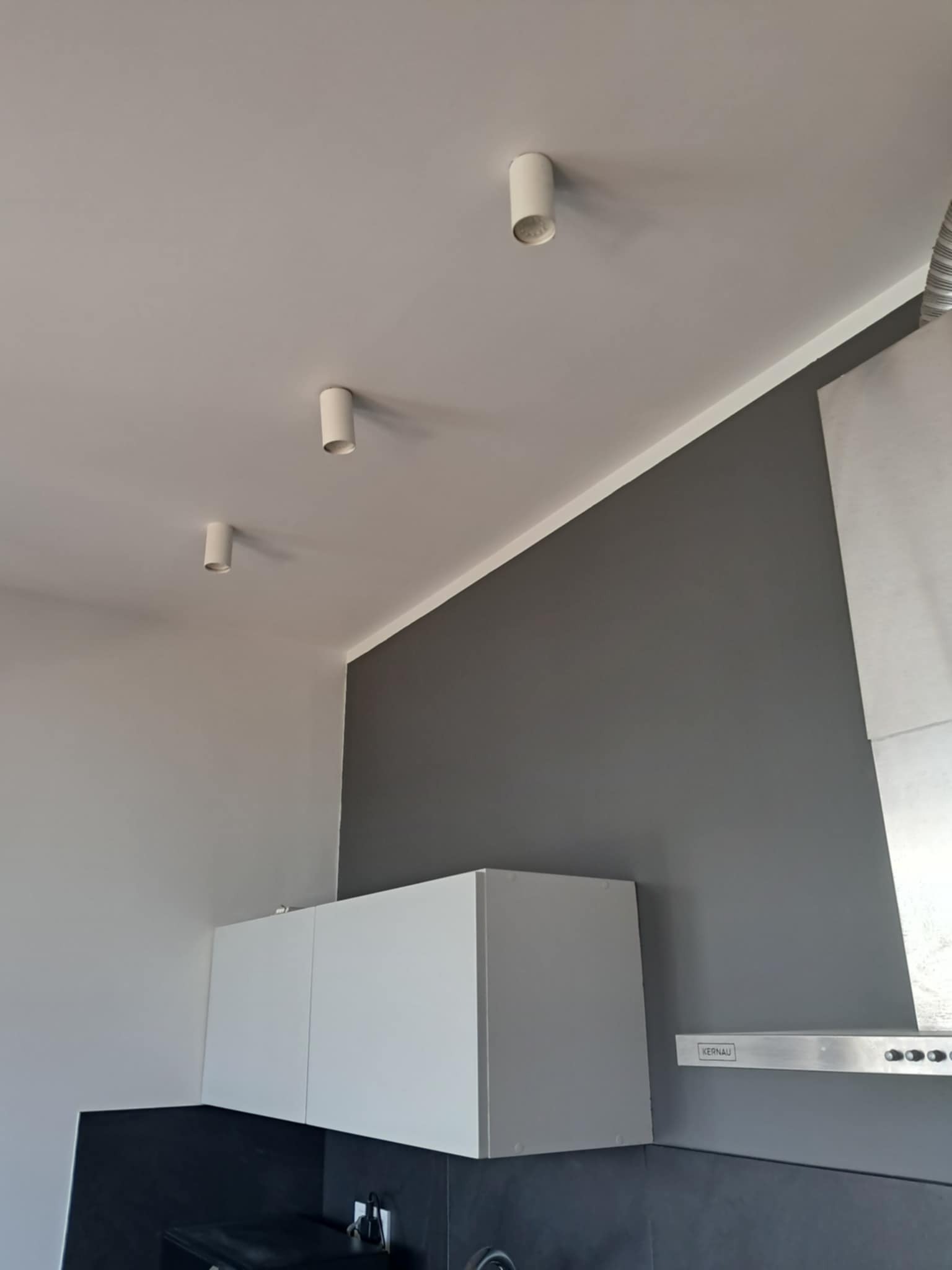 Nowoczesna kuchnia po malowaniu: szara ściana kontrastuje z białymi szafkami i sufitowymi lampami tubami. Widoczny fragment okapu kuchennego.