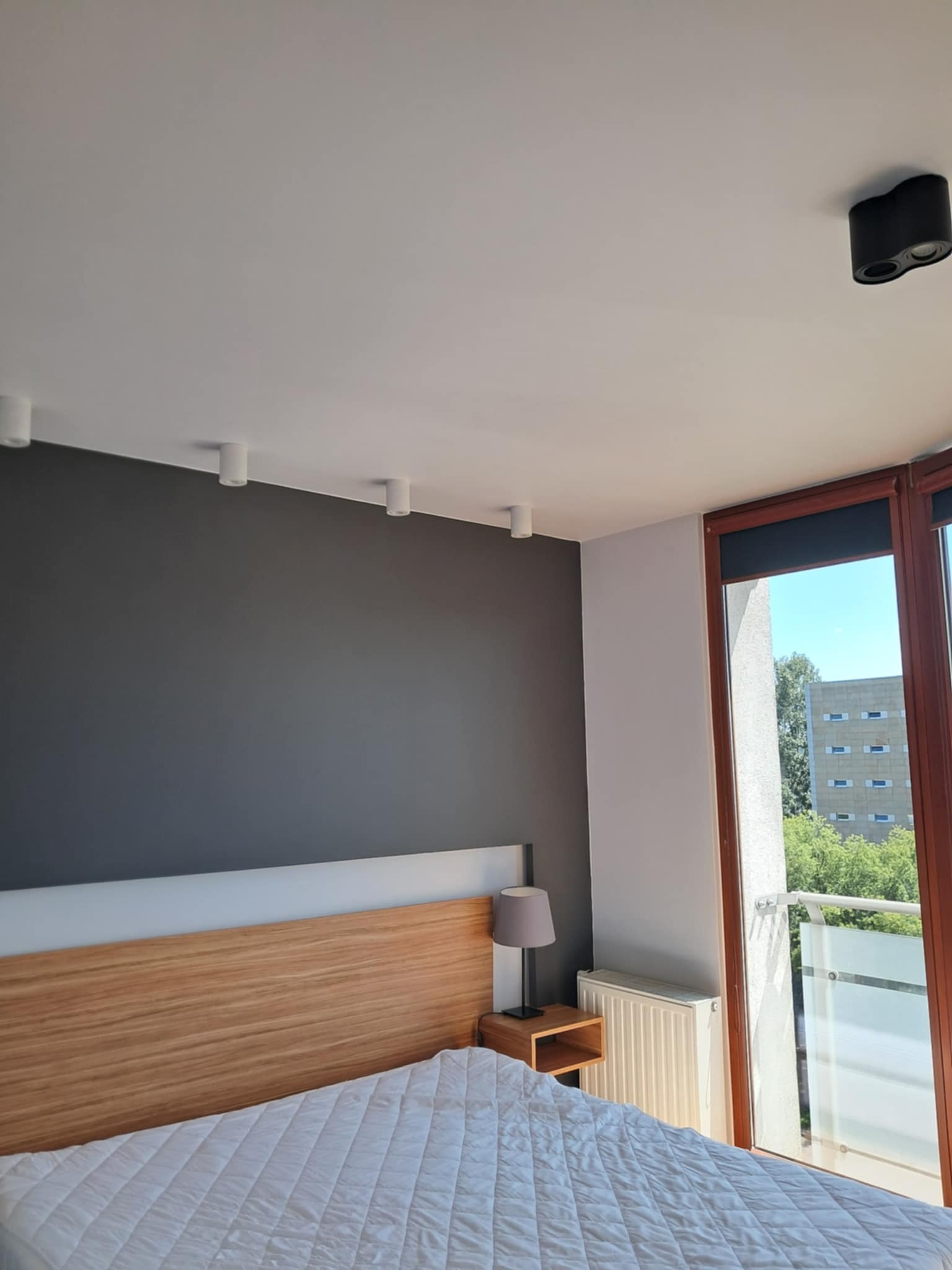 Jasne wnętrze sypialni z szarą ścianą, drewnianym zagłówkiem i oknem balkonowym z widokiem na zieleń. Oświetlenie sufitowe punktowe.
