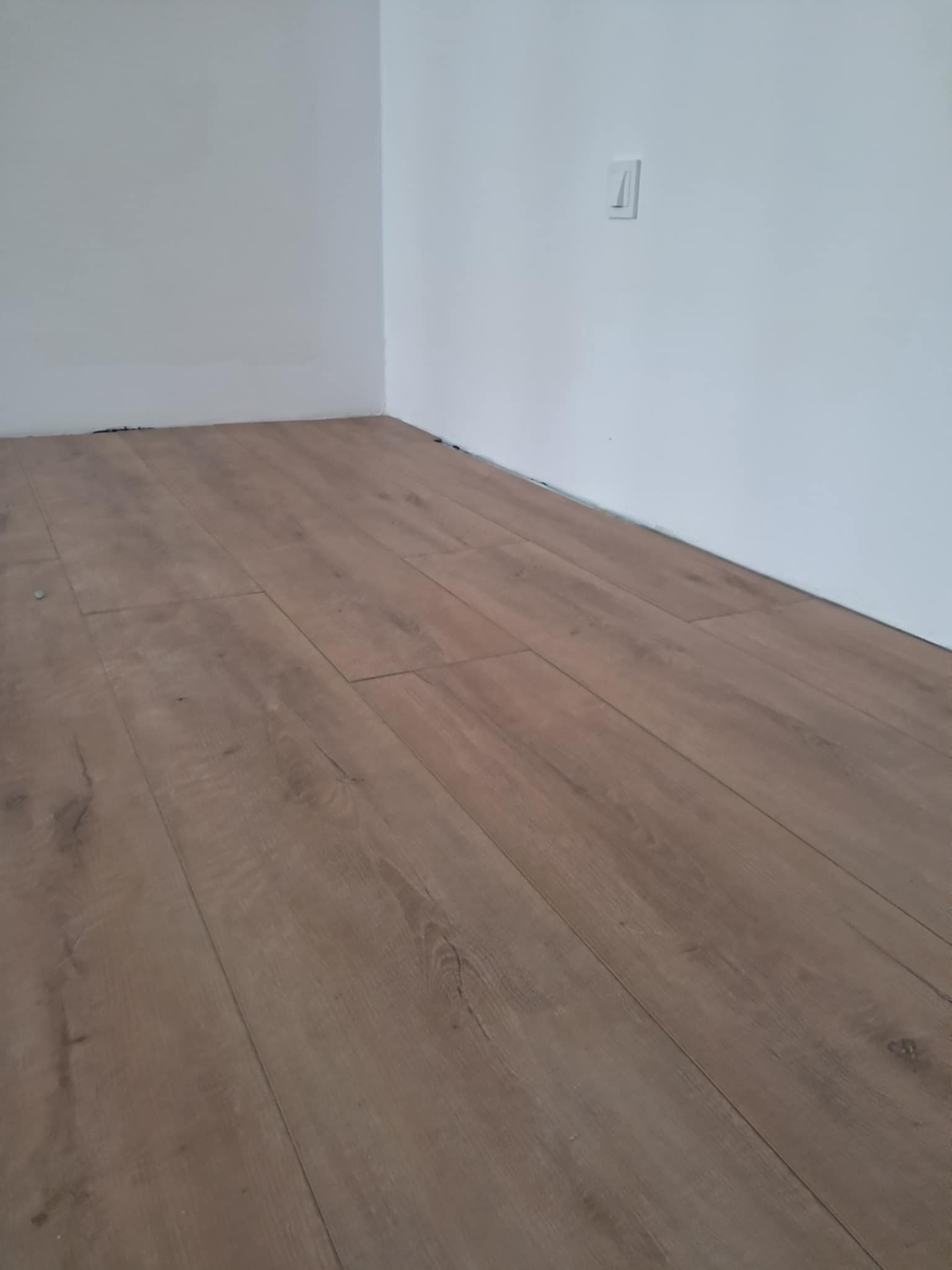 Ułożone panele podłogowe w rogu pomieszczenia, widoczne łączenia i faktura drewna, ściany pomalowane na biało, widoczny włącznik elektryczny.