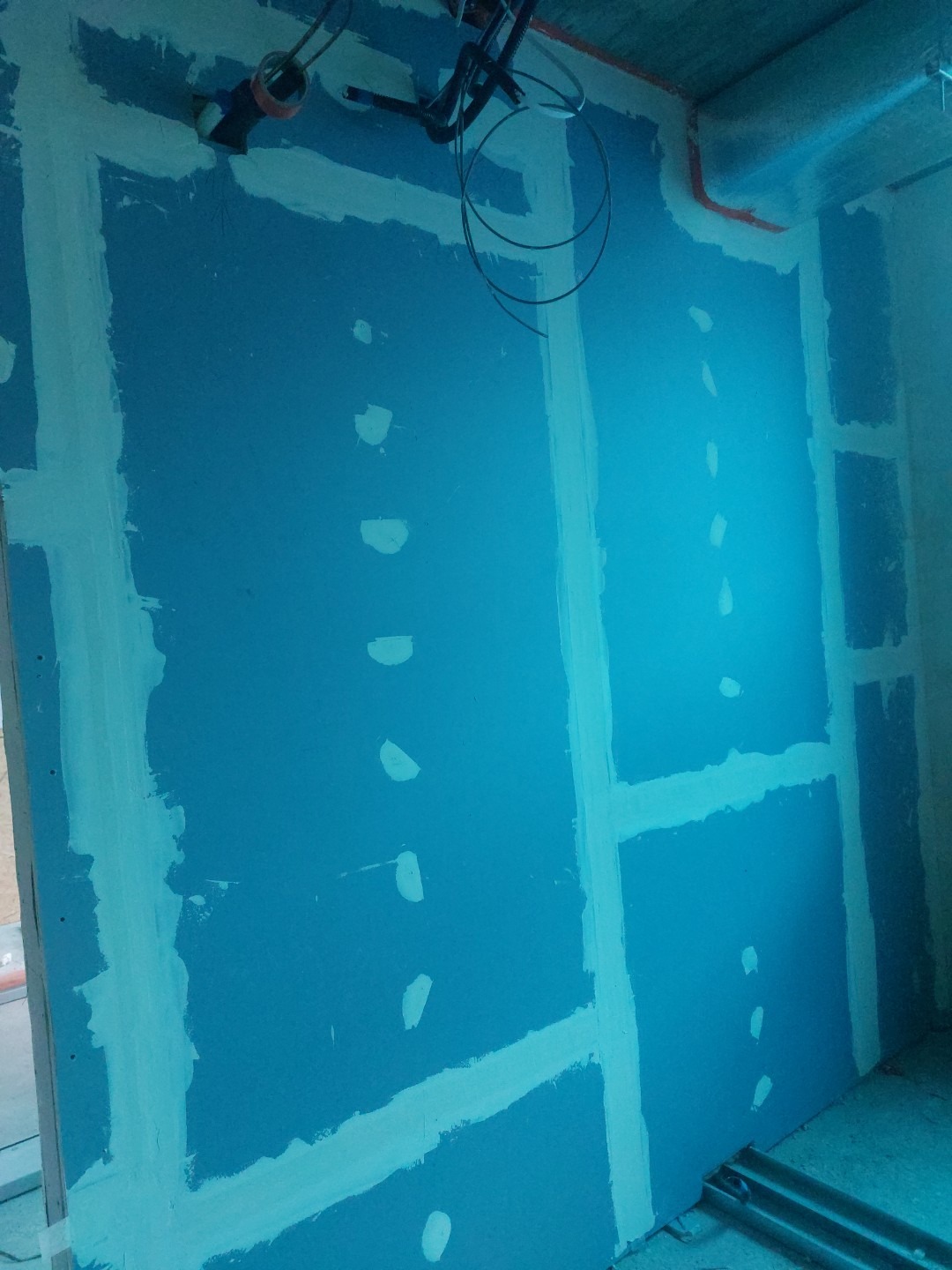 Niebieska ściana z płyt kartonowo-gipsowych, szpachlowane łączenia, widoczne przewody elektryczne, sufit z elementami instalacji.