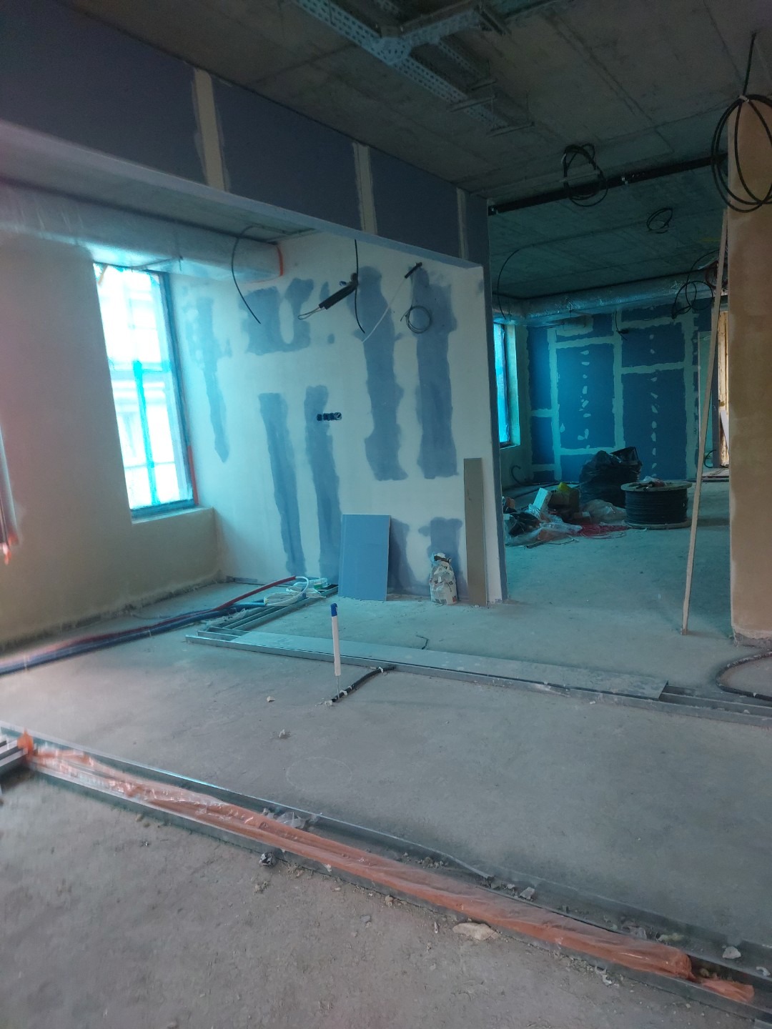 Wnętrze w trakcie prac wykończeniowych: ściany z płyt gips-karton, widoczne instalacje, surowy beton na podłodze. Okno z niebieską folią zabezpieczającą.