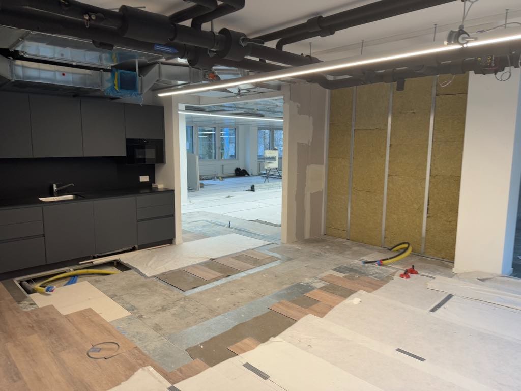 Wnętrze biura w trakcie remontu: montaż płyty karton-gips, układanie paneli podłogowych i instalacja nowej zabudowy kuchennej w odcieniach szarości. Widoczne rury wentylacyjne pod sufitem.