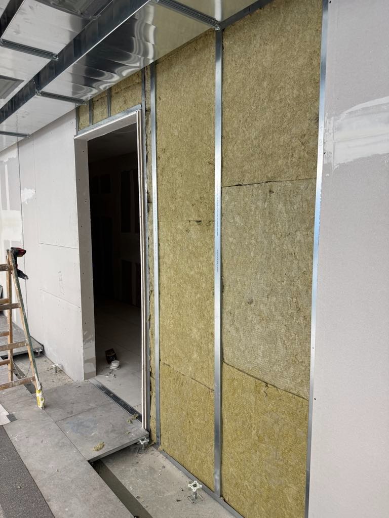 Montaż izolacji z wełny mineralnej w metalowym stelażu ściany działowej, widoczne płyty gipsowo-kartonowe i drabina. Wnętrze w trakcie prac wykończeniowych.