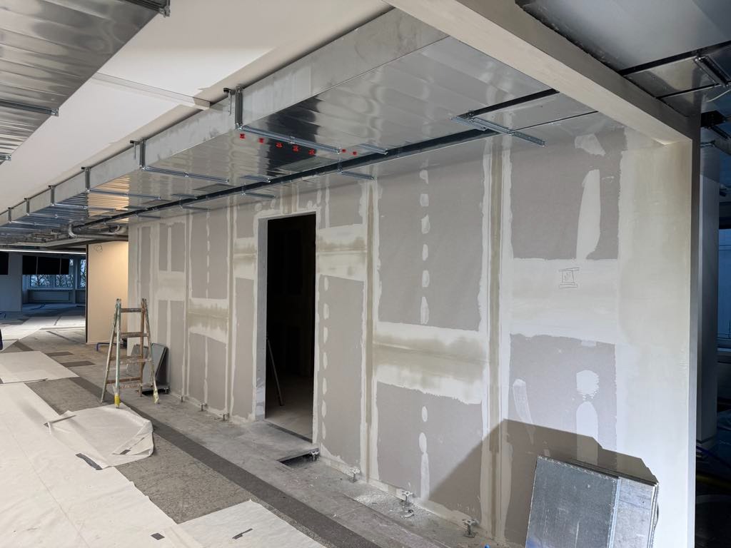 Wnętrze biura w trakcie remontu: ściany z płyt karton-gips, widoczne profile sufitowe i drabina. Surowy stan wykończenia, przygotowanie do dalszych prac.