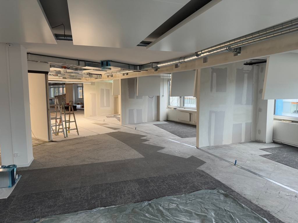 Biuro w trakcie remontu: widoczne ściany z płyt karton-gips, metalowe rury pod sufitem, drabina i zabezpieczona podłoga. Etap prac wykończeniowych, surowy stan.