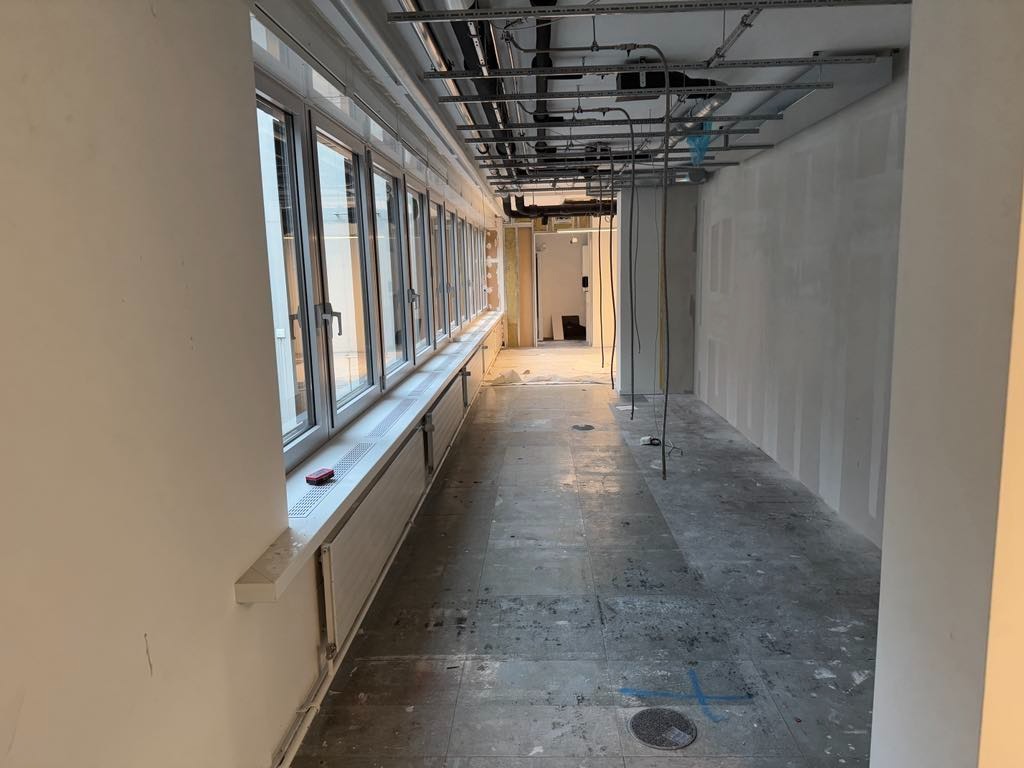 Wnętrze biura w trakcie remontu: ściany z płyt karton-gips, okna wzdłuż ściany, odsłonięta instalacja pod sufitem i surowa podłoga przed wykończeniem.