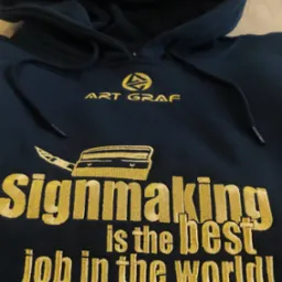Czarna bluza z kapturem, złoty haft: logo Art Graf, grafika noża introligatorskiego i napis 'Signmaking is the best job in the world!'.