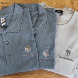 Ewelina Mularczyk Studio Personalizacji Odzieży - Jasnoniebieska koszulka polo i szare t-shirty z identycznym, szarym haftem logo 'Isosolutions' ułożone na drewnianym stole. Widok z góry.