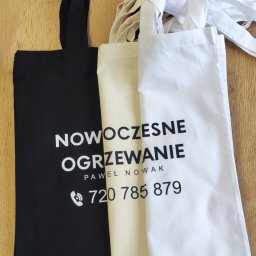 Ewelina Mularczyk Studio Personalizacji Odzieży - Trzy torby bawełniane z nadrukiem reklamowym: czarna z napisem 'Nowoczesne Ogrzewanie Paweł Nowak 720 785 879', beżowa i biała, leżące na drewnianym blacie.