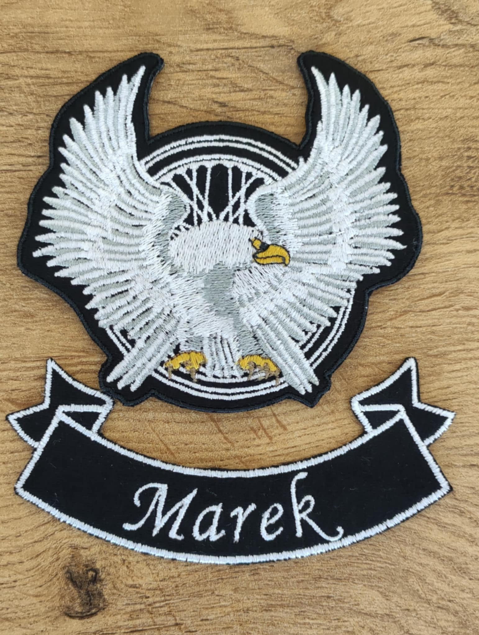Czarno-biała naszywka haftowana z motywem orła w kole i banerem z napisem 'Marek' na drewnianym tle.