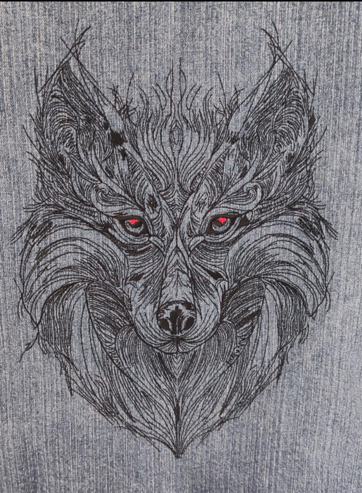 Szczegółowe haftowane przedstawienie głowy wilka z czerwonymi oczami na denimowym tle.