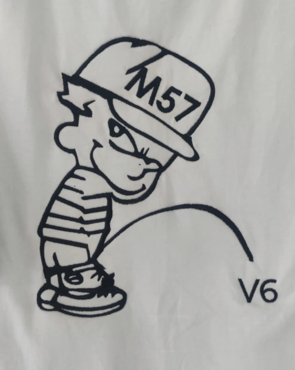 Biała koszulka z czarnym nadrukiem przedstawiającym kreskówkową postać chłopca w czapce z napisem M57, sikającego na napis V6.