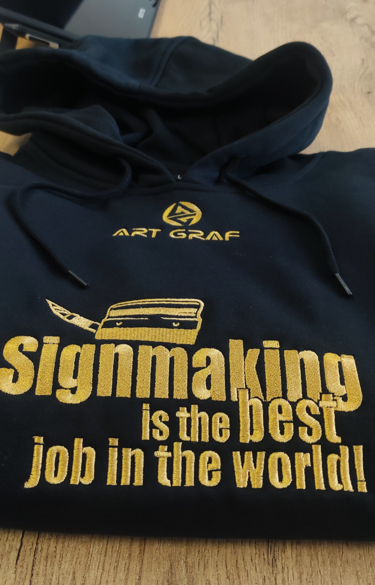 Czarna bluza z kapturem, złoty haft: logo Art Graf, grafika noża introligatorskiego i napis 'Signmaking is the best job in the world!'.