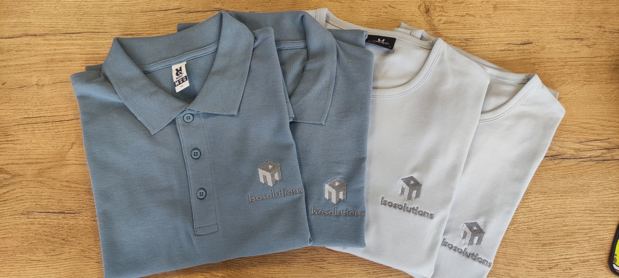 Jasnoniebieska koszulka polo i szare t-shirty z identycznym, szarym haftem logo 'Isosolutions' ułożone na drewnianym stole. Widok z góry.