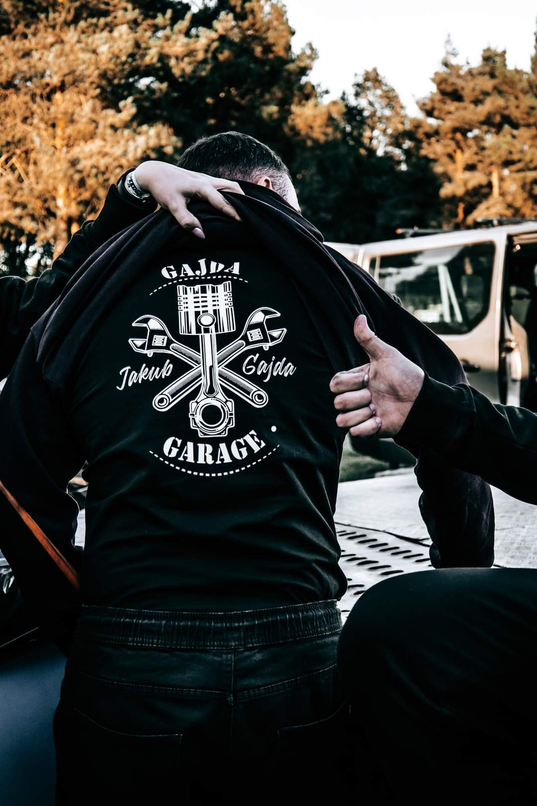 Czarna bluza z białym nadrukiem: tłok, klucze i napis 'Gajda Garage'. Osoba zakłada bluzę, widok z tyłu, na zewnątrz, w tle drzewa i samochód.