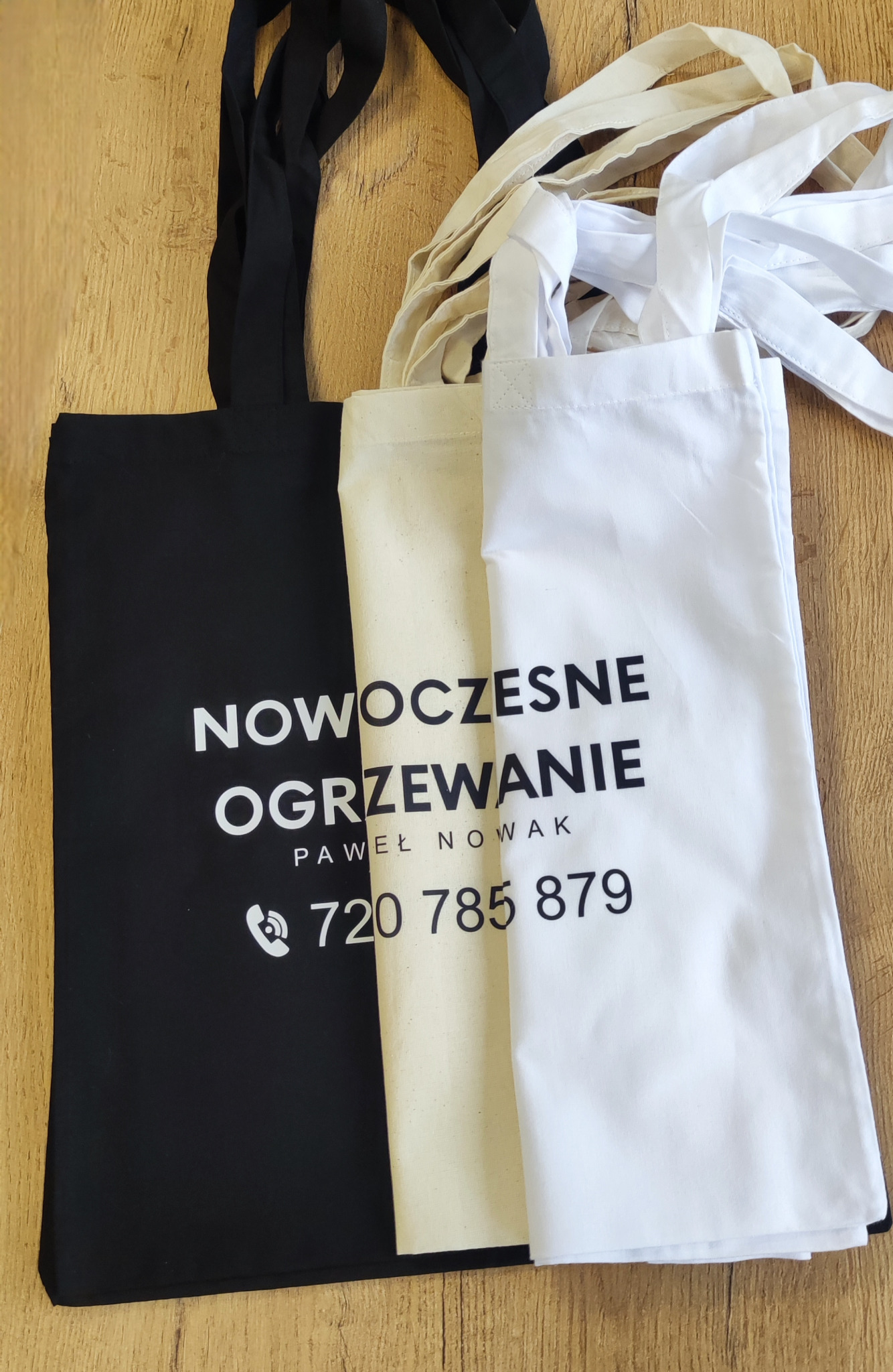Trzy torby bawełniane z nadrukiem reklamowym: czarna z napisem 'Nowoczesne Ogrzewanie Paweł Nowak 720 785 879', beżowa i biała, leżące na drewnianym blacie.