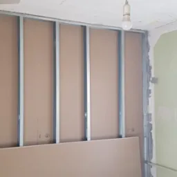 Montaż stelaża z profili metalowych i płyt gipsowo-kartonowych na ścianie w trakcie prac wykończeniowych, widoczna surowa ściana w tle i żarówka na suficie.