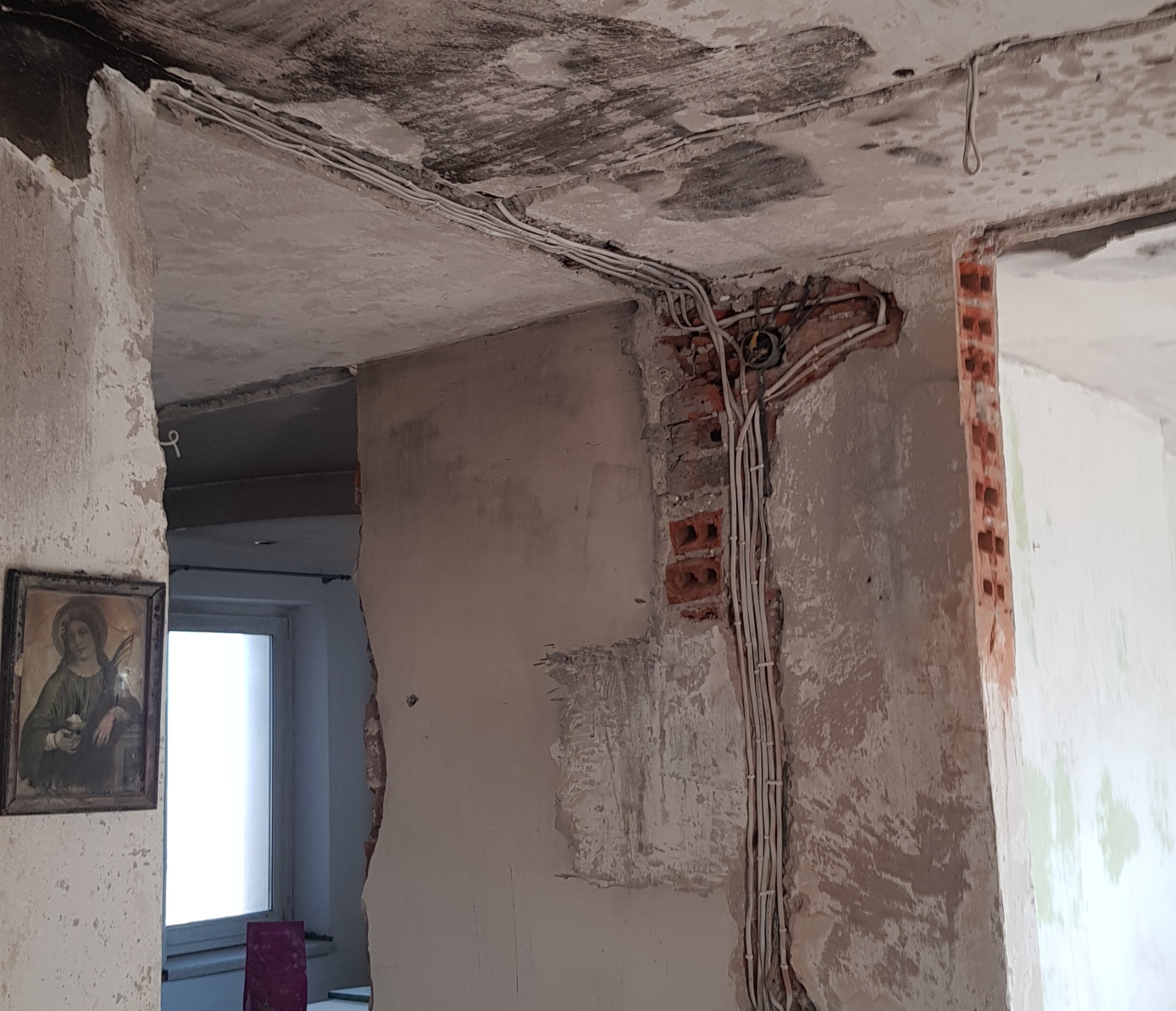 Pokój w trakcie prac przygotowawczych do wykończenia: odsłonięta cegła, poprowadzone kable elektryczne, ściany z resztkami starej farby i tynku, obraz religijny w ramce.