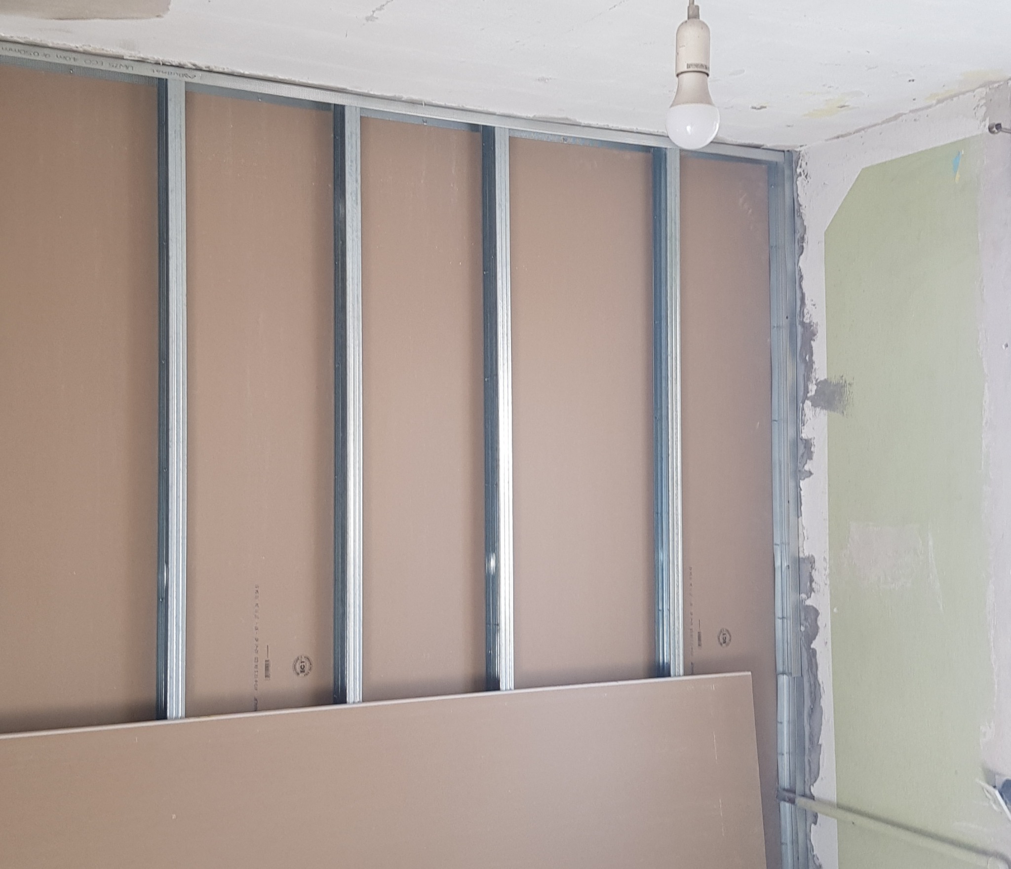 Montaż stelaża z profili metalowych i płyt gipsowo-kartonowych na ścianie w trakcie prac wykończeniowych, widoczna surowa ściana w tle i żarówka na suficie.